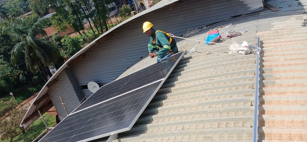 Worker in yellow hard hat installing dark shingles on residential roof with completed brick section visible